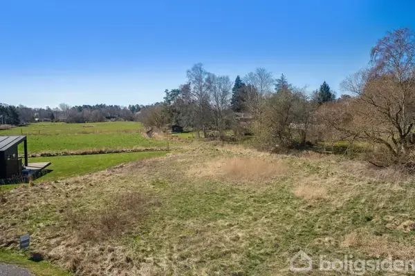 En åben græsmark med enkelte buske og træer omkranser et lille, moderne hus til venstre. Blå himmel og skov i baggrunden. Ingen synlig tekst.