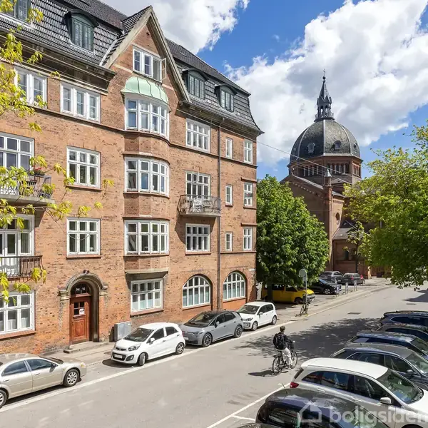 En stor, rød murstensbygning med hvide vinduer står ved en gade, hvor biler er parkeret. En person cykler forbi. I baggrunden ses en kirke med en stor kuppel.