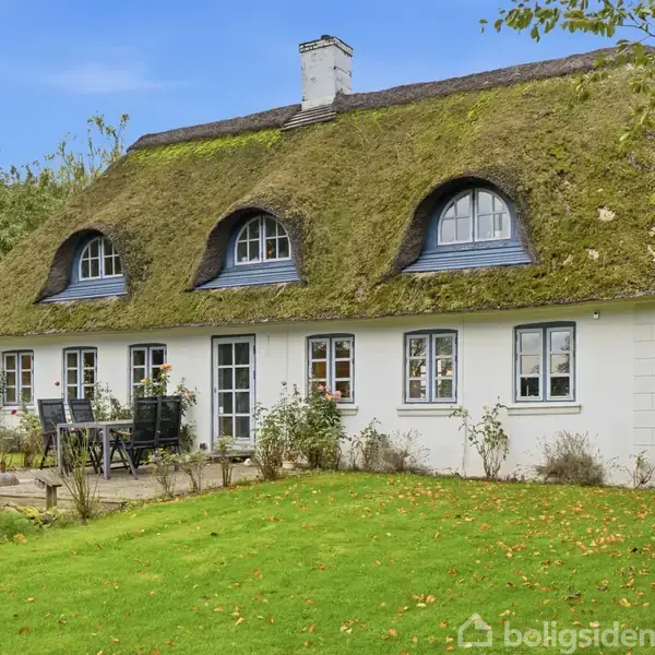 Et hvidt bindingsværkshus med stråtag står på en grøn græsplæne. Foran huset er en terrasse med havemøbler. Omgivet af træer og buske i en landlig have.