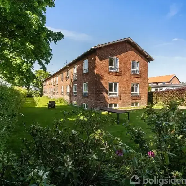 Rødt murstenshus med flere vinduer står stille. Det er omgivet af en grøn have med buske og træer under en klar blå himmel.