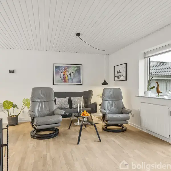 Stue med to grå lænestole foran grå sofa, to glasborde med lys og appelsiner, plankegulv, hvide vægge, stort vindue, plante, og vægkunst i lyse omgivelser.