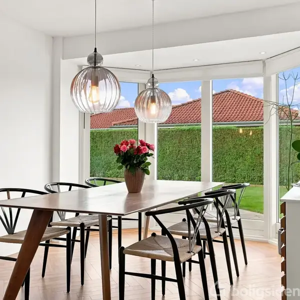 Spisebord med blomsterbuket står i spisestue. Modern stil med seks stole. Store vinduer viser have med hæk og røde hustage. To hængelamper lyser rummet.