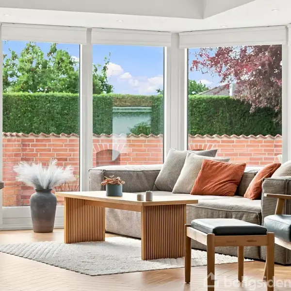 Sofa med farvede puder står ved store vinduer, der lukker naturligt lys ind. Foran er et sofabord og en skammel. Udenfor ses en murstensvæg og buske.