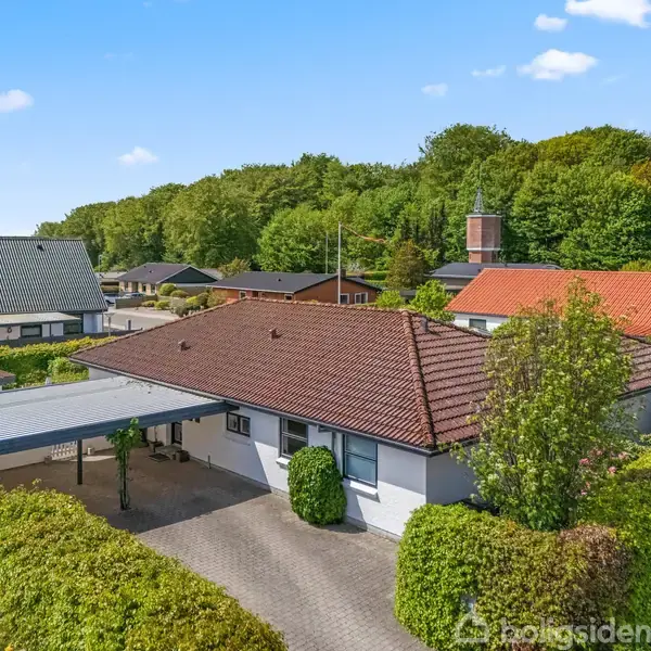 Et hus med rødt tegltag og carport står stille omgivet af hække og træer i en forstadslignende kvarter med flere lignende boliger og en skov i baggrunden.