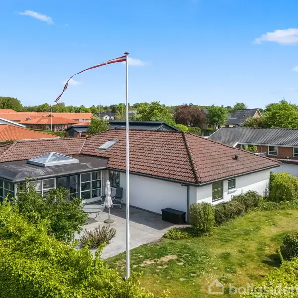 Et rødt tegltagshus stående i en frodig have omgivet af grønne buske og træer; en flagstang med et flag vifter i vinden foran huset.