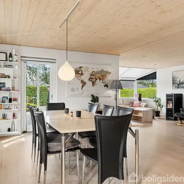Spisebord med sorte stole står på trægulv. En stor lampe hænger over bordet. Reol med bøger langs væg. Stue med sofa og brændeovn i baggrunden. Verdenskort på væggen.