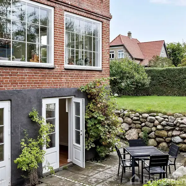 Rød murstensbygning med hvide vinduesrammer, en åben havedør omgivet af klatreplanter. I baggrunden en græsplæne afgrænset af en stensætning og en velplejet hæk.
