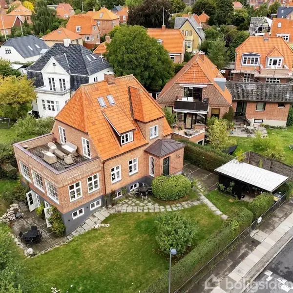 Et rødt murstenshus med teglstenstag står i en frodig have. Omkring huset er der flere lignende boliger. En asfalteret vej løber langs området, kantet af grønne buske.