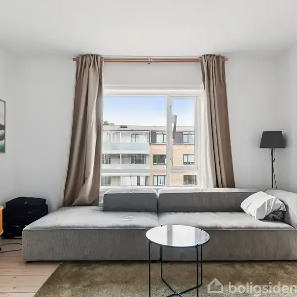 Grå sofa står foran et stort vindue med beige gardiner, omgivet af en fladskærmstv og en gulvlampe. Bygninger ses udenfor. Sorte og hvide puder ligger på sofaen.