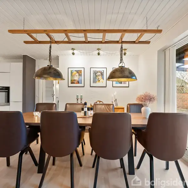 En lang spisebord med otte brune stole står under to store loftslamper. I baggrunden ses et moderne køkken og tre kunstværker på væggen. Store vinduer åbner ud til terrassen.