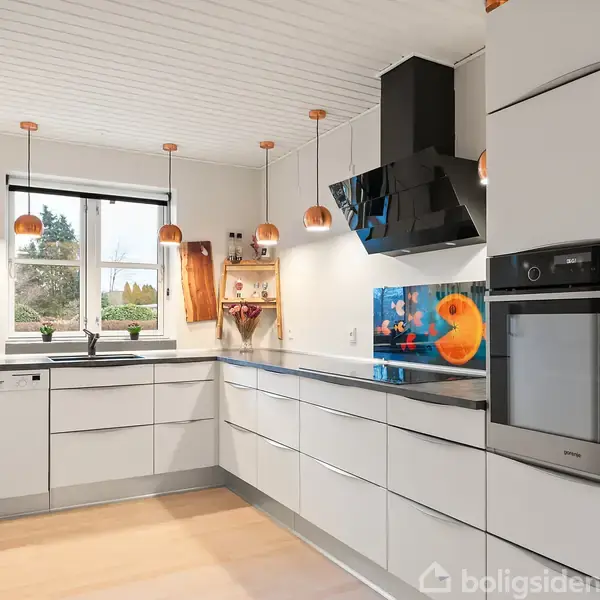 Et moderne køkken med hvide skabe og sorte bordplader. Der hænger knive ved siden af vasken. Et farverigt akvariebillede er på væggen. Store vinduer giver naturligt lys.