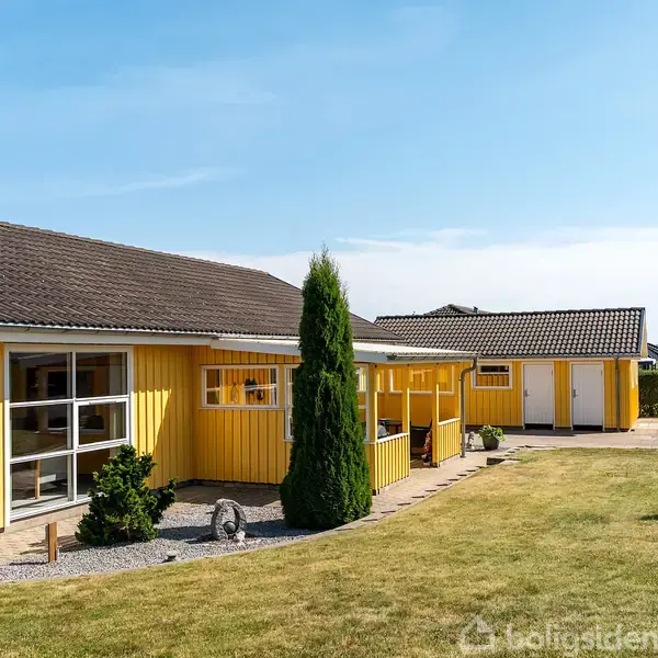 Gult hus med store vinduer står stille i en velplejet have med grøn plæne. Små buske og et træ omgiver huset, mens himlen er klar og blå.