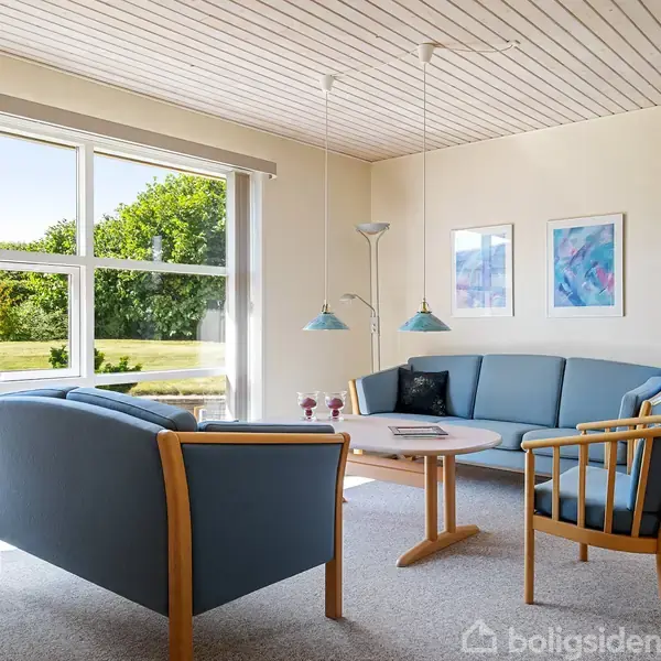 En stue med blå sofa og stole omkring et rundt bord. To lamper hænger over bordet. Store vinduer viser en have med græs og træer udenfor.