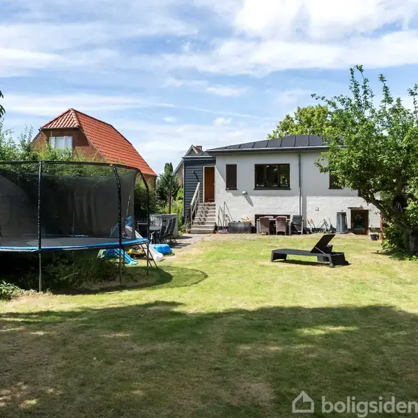 En villa med hvid facade og fladt tag står for enden af en græsplæne. En trampolin er placeret til venstre, og et træ med grønne blade ses til højre.