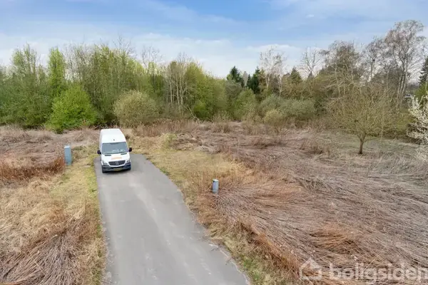 En hvid varevogn holder på en smal asfalteret vej. Omgivet af tørt græs og en skovbevokset baggrund under en klar himmel.