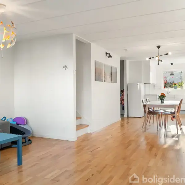 En moderne, hvid to-etagers rækkehusbygning har en parkeret bil i carporten. Omgivet af grønne buske og en flisebelagt indkørsel. Et skilt viser tallet "15".