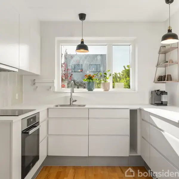 Et moderne køkken med hvide skabe, en indbygget ovn og lys træbund. To lamper hænger over bordpladen. Et vindue viser planter udenfor, og planter står også i vindueskarmen.