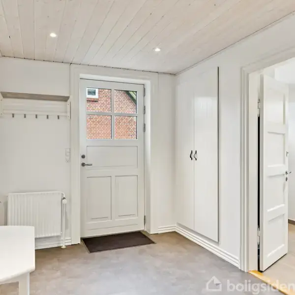 Dør står åben i en lys gang med hvidt loft. En plante står på bord til venstre, garderobeskab til højre. Gulv er gråbrunt, murstensbygning ses gennem vindue.