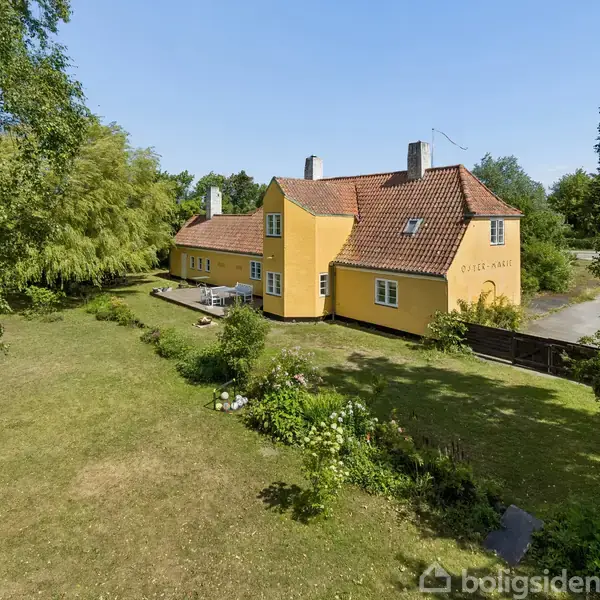 En gul, to-etagers bygning med tegltag står i en frodig have med træer og græsplæne. Der er teksten "ODEL HUS" på huset, omgivet af natur.