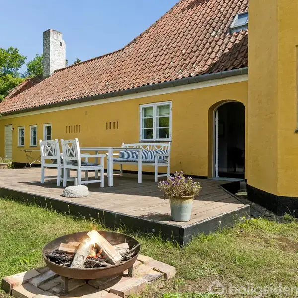 Gulmalet hus med rødt tegltag og hvid rammevinduer. En flisebelagt terrasse med havemøbler står foran huset. En bålplads er tændt på græsplænen ved siden af.