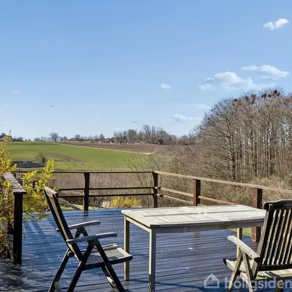 Træbord og to stole står på en veranda. Omgivet af landlig natur med marker og træer under en klar blå himmel.