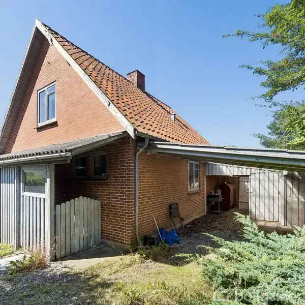 Et rødt murstenshus står stille med en skråtag under en klar blå himmel. Omringet af grøn vegetation og en lille grussti, ses et skur ved siden af.