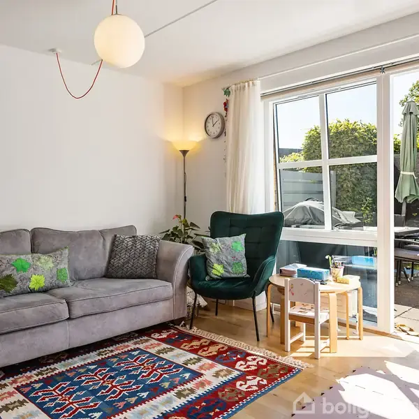 Gråt sofaarrangement med farverige puder står i en stue. Ved siden af er en grøn stol og små borde. Glasdøre i baggrunden fører ud til en terrasse og have med trampolin.