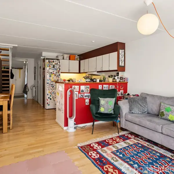 Stue med sofa og lænestol, vendt mod et køkken med rød bar. Spisebord til venstre og farverige tæpper på gulvet. Køkkenet har hvide skabe og mange køleskabsmagneter.