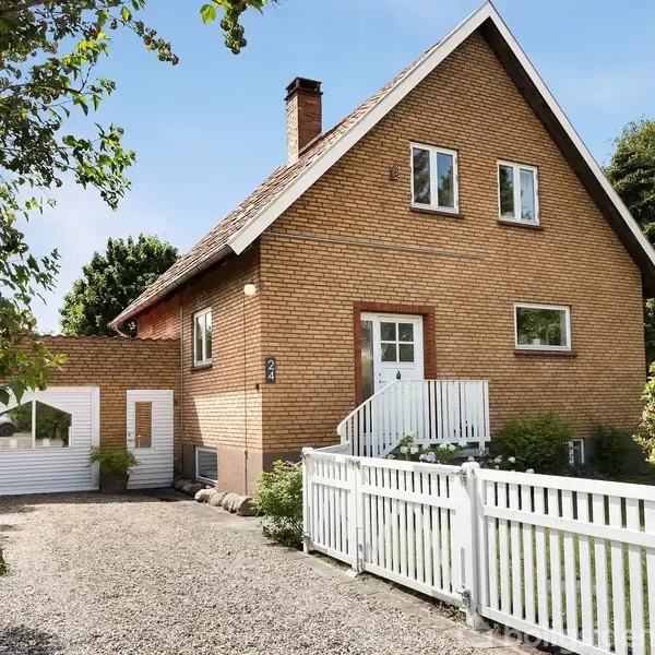 Et murstenshus med hvidt stakit og garage står i en grøn have. Huset har en trappe foran døren og vinduer i tagetagen. Sollys skinner på facaden.