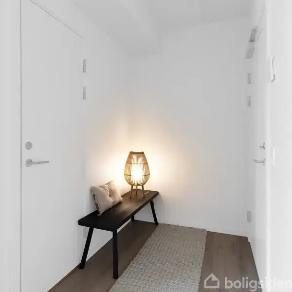 Bænk med puder står mod en hvid væg i en gang. En tændt lampe på bænken skaber en varm glød. Rummet har lyse vægge og et gulvtæppe.