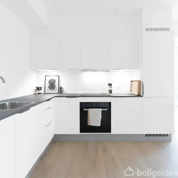 Et moderne, minimalistisk køkken med hvide skabe, sort bordplade og indbygget ovn. En stålvask med en enkel hane er ved siden af ovnen. Der er en plante og stol i baggrunden.
