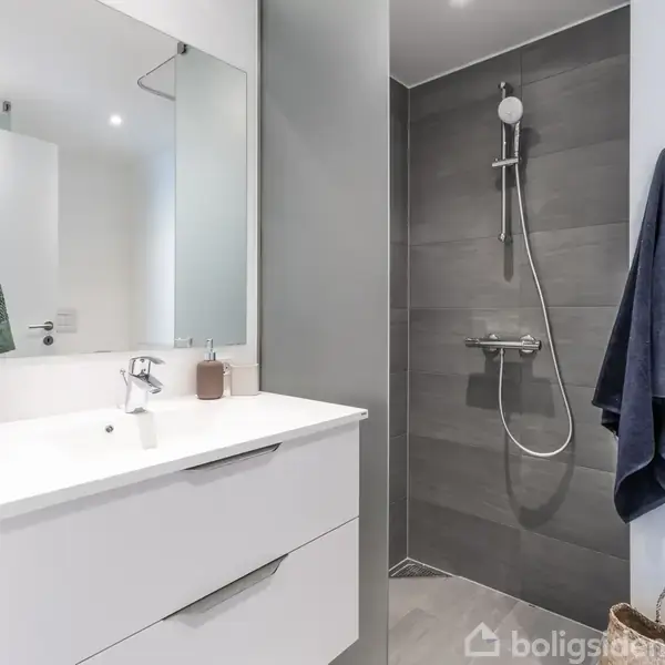 Håndvask med spejl hænger i et moderne badeværelse. Bruseren er monteret på en mørk væg til højre. Hvide og blå håndklæder hænger på væggen.