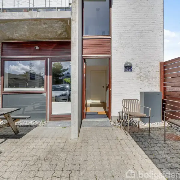 En åbnet hoveddør fører ind til et hus, omgivet af en belagt terrasse med et træbord og to terrassestole. Røde træhegn omkranser området.