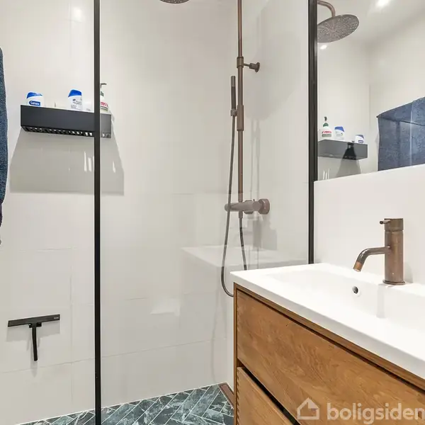 Bruseområde med glasvæg og moderne brusehoved. Ved siden af er en vask med brun vandhane og træskab. Hylder med produkter er monteret på væggen. Ingen tekst fundet.