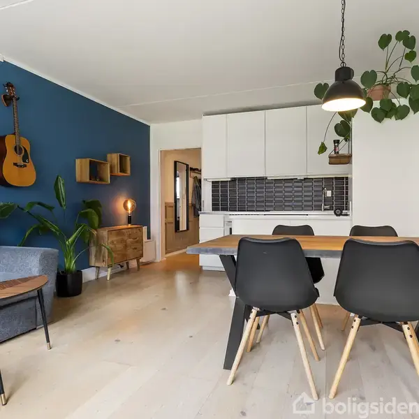 En moderne stue med blåmalet væg, hvorpå en guitar hænger. En grå sofa med et sofabord foran. Et spisebord med sorte stole står nær et åbent køkken med hvide skabe. Planter dekorerer rummet.