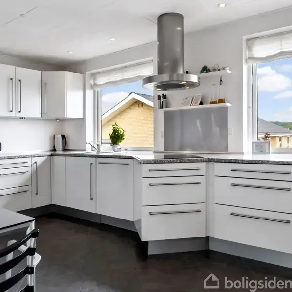 Et moderne køkken med hvide skabe og stålgreb har en ovn indbygget. En emhætte hænger over komfuret. Store vinduer giver udsigt til en gårdsplads og landskab.