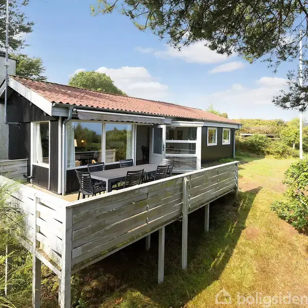 Et sommerhus med træterrasse er omkranset af grønne træer og buske. En lang række mørke havemøbler står på terrassen, der strækker sig ud over en frodig have.