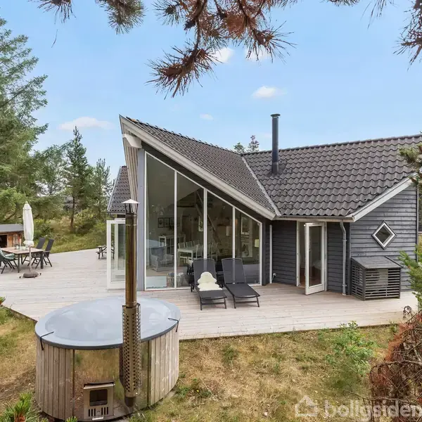 Moderne sommerhus med skarpt vinklet tag står på træterrasse med loungestole og udebruser som handling. Omgivet af skov, skaber huset en fredelig, naturskøn atmosfære.
