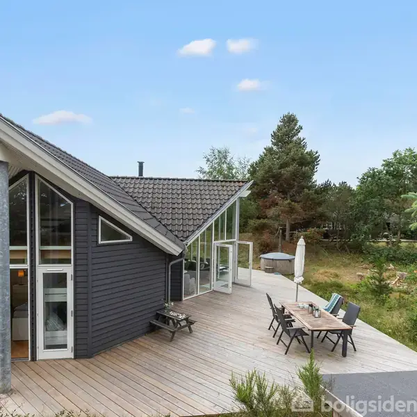 Sortmalet sommerhus med store vinduer, stående på en træterrasse. Der er havemøbler og en parasol på terrassen. Omgivet af grønne træer og en klar, blå himmel.