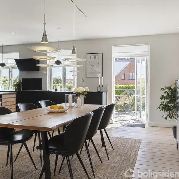 En spisebord med sorte stole står i et lyst køkkenalrum. Bordet er pyntet med frugtskål og blomster. Baggrunden viser køkkenskabe og en udgang til terrassen med udsigt til huset.