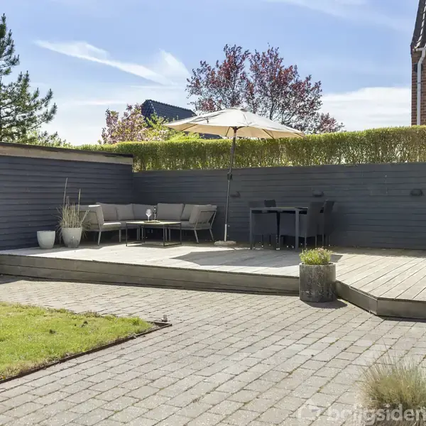 En flisebelagt terrasse med en hjørnesofa og parasol står på en hævet platform. Til højre ses en grill i et indhegnet haveområde omkranset af hækketræer og en murstensbygning.