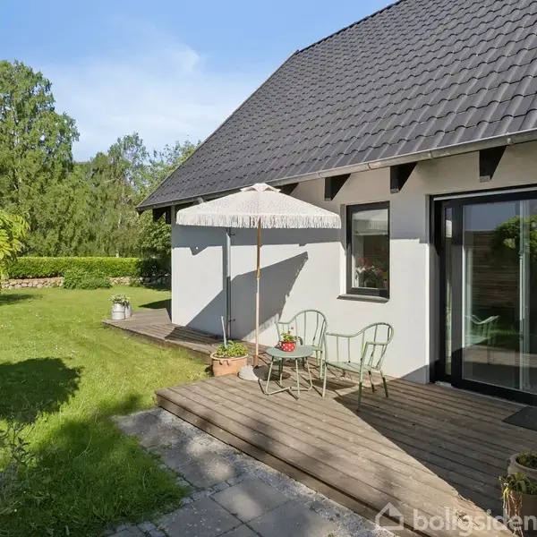 Huset har en terrasse med en parasol og to stole. Det står i en grøn have med frodige træer og buske, belyst af sollys.