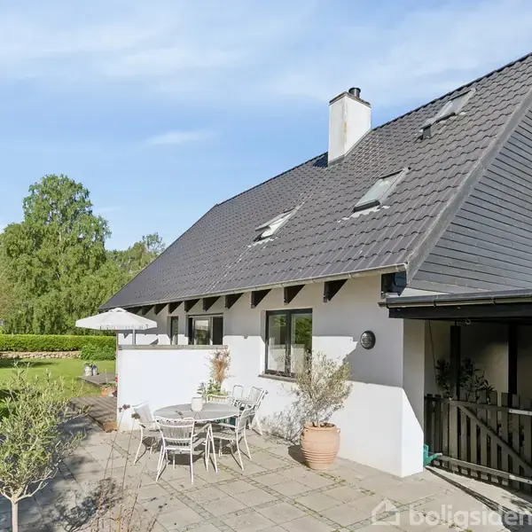 Huset har en moderniseret tagkonstruktion med tre vinduer, der under en skorstensrør. Foran er der en terrasse med bord, stole og parasol, omgivet af en frodig have.
