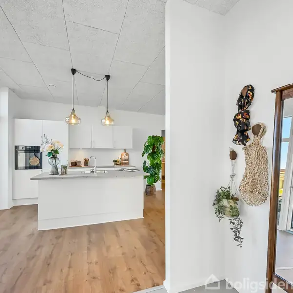 Et moderne køkken med hvidt interiør og to hængende lamper, står i det lyse rum med store vinduer, trægulv og udsigt til terrasseområdet udenfor.