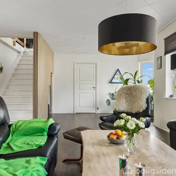 Sorte lædersofaer står i en stue med et træbord dekoreret med blomster og frugtskål. En trappe fører til en overetage, omgivet af planter og moderne belysning.