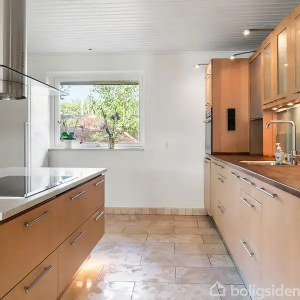 Et moderne køkken med lyst træinteriør, hvor en kogeplade med emhætte er centralt placeret. Der er store skabe langs væggen, og et vindue giver udsigt til en grøn have.