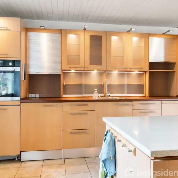 Et køkken med lyse træskabe og en indbygget ovn. Skabet er åbent med hylder, og der er en stor, lys køkkenø i forgrunden. Flisegulv omgiver området.