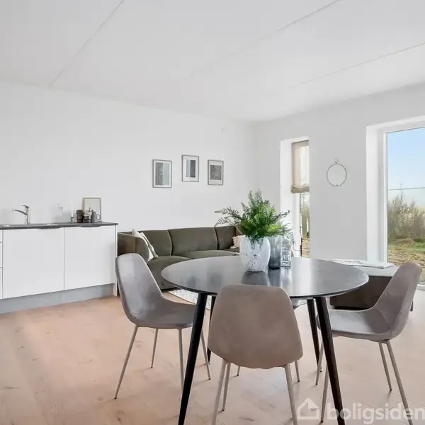 Rundt spisebord står i en lys stue med fire stole omkring det. Køkkenbordet er til venstre, mens sofa og kunstværker ses i baggrunden. Store vinduer giver udsigt til naturlig vegetation.