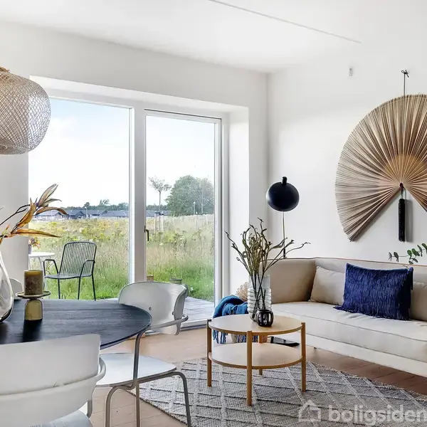 Stue med moderne indretning, hvor en sofa med blå puder står ved siden af et rundt sofabord og kunst på væggen. Store vinduer åbner op til en grøn have.