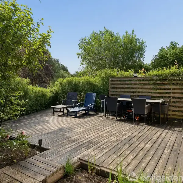 Terrasse med træbrædder har liggestole og spisebord med stole. Det hele er omgivet af buske og træer, hvilket skaber en hyggelig udendørs atmosfære.